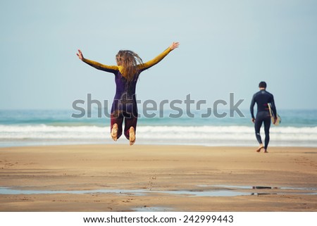Young surfer girl jump having fun while her friend walk to the ocean ready to surfing