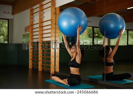 Young athletic woman holding up balance ball and sitting on the mat during her fitness training in gym