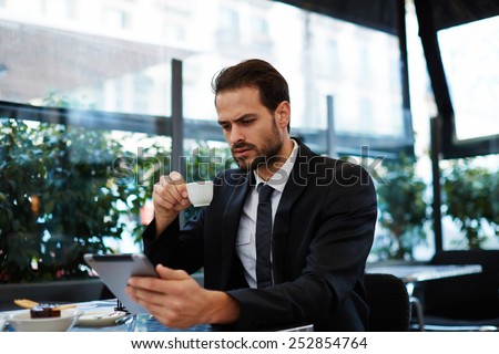 Young business man having a breakfast sitting on beautiful terrace with plants, handsome man in suit read news holding digital tablet pc in the hands