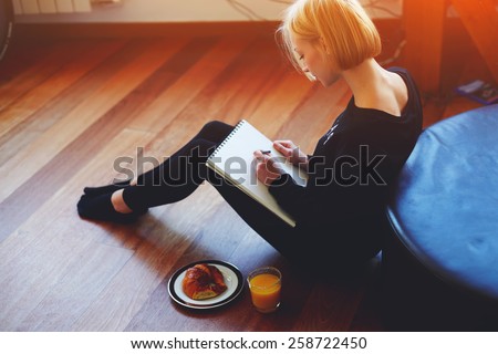 Beautiful young woman writing something in the note pad while sitting on the floor at living room, charming female studying doing homework at home, girl writes diary at her breakfast, flare light