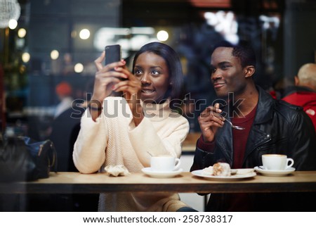 Two dark skinned stylish friends having breakfast in cafe, attractive black woman taking the picture with digital camera on smart phone sitting in beautiful coffee shop, view through cafe window