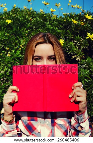 Portrait of charming young woman with pink book held up close to her face, cute female covering half face with a book while standing on spring green hedge with flowers background, filtered image