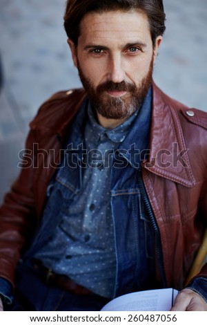 Portrait of mature stylish man holding modern magazine while sitting on the terrace of coffee shop, stylish hipster with beard focused look to the camera