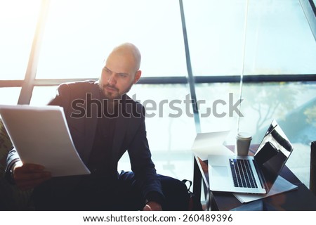 Portrait of elegant business man looking over some paperwork sitting next to big window in office interior, male executive studies documents or papers during his coffee time, flare sun light