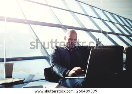 Shot of a handsome businessman typing on computer keyboard and reviewing documents sitting in light modern office interior, managing director examining paperwork in coffee shop, flare sun light