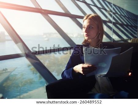 Portrait of young elegant woman sitting in modern office interior holding papers and pensively gazing out of the window, filtered image with flare sun light from the window