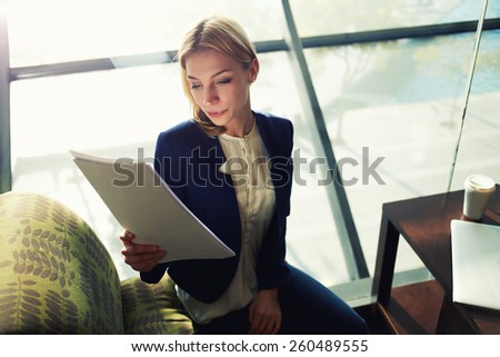 Portrait of young blond hair business woman examining paperwork in bight light office interior sitting next to the window, attractive business woman read some documents before meeting
