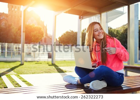 Charming blonde female student pointing to open laptop while looking for someone at campus, happy female teenager having fun while sitting on the bench with open computer outdoors, flare bright sun