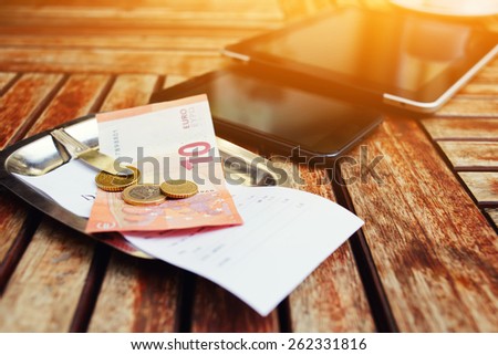 Mock-up with bill check and money, smart phone and touch screen digital tablet on the wooden desk, gadgets with empty blank screen, work break of businessman, flare sun light
