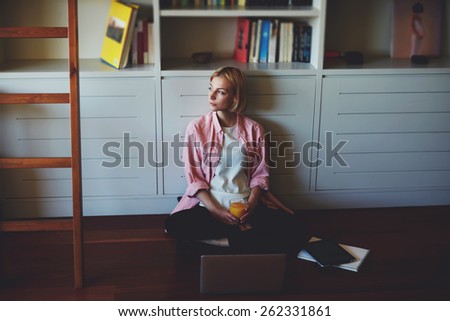 Gorgeous young woman sitting at bookshelf on the floor with open laptop computer while she works with comfort from home, young business woman drink orange juice while having breakfast, young student