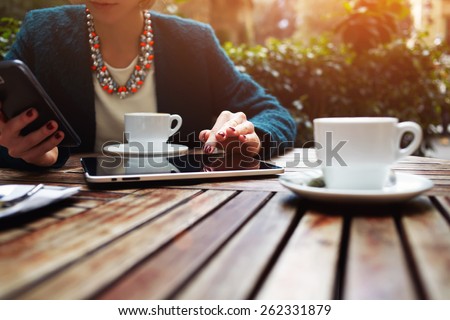 Cup of coffee on the foreground with elegant young woman using busy touch screen tablet at the coffee shop wooden table, work break of business people, flare sun light
