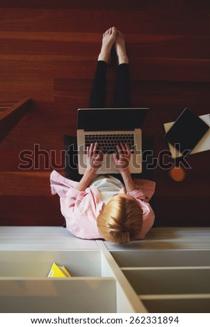 Top view young female freelancer busy working on laptop computer while leaning on home bookshelf sitting on the floor, charming business woman working from home while having breakfast, young student