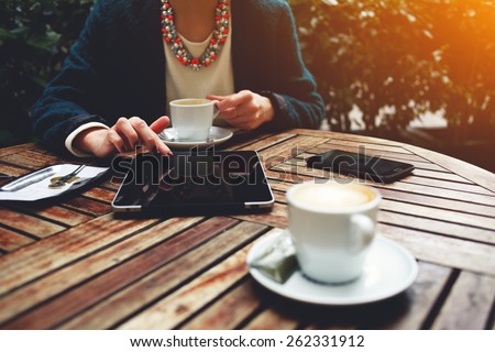 Cup of coffee on the foreground with elegant woman using busy touch screen tablet and drinking cappuccino at the coffee shop, bill check and mobile phone near, flare sun light