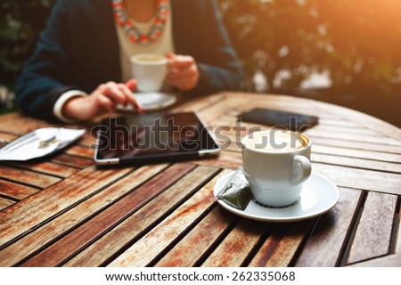 Cup of coffee on the foreground with elegant woman using busy touch screen tablet at the coffee shop wooden table, bill check and mobile phone near, flare sun light