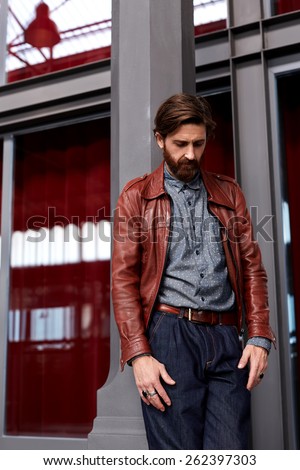 Portrait of stylish dressed man leaning on interior building pillar while posing indoors, fashionable mature man with beard on beautiful glassy background