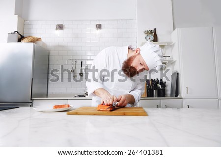 Portrait of professional and experienced chef cook in uniform diligently cutting salmon fish on fillets with knife on a board, male chef preparing gourmet meal of seafood in modern kitchen