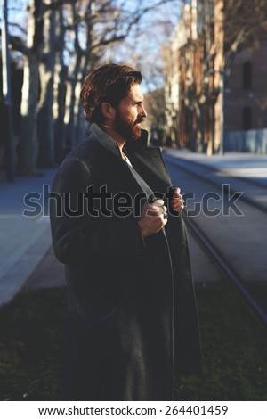 Portrait of fashionable well dressed man with beard posing outdoors looking away, confident and focused mature man in coat standing outside at sunny evening, elegant fashion model