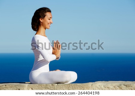 Side shot of smiling beautiful young woman practicing yoga on a sunny day with amazing sea horizon on background, woman seeking enlightenment through meditation, relaxed girl performing yoga routine