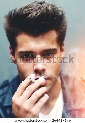 Close up portrait of handsome man exhaling cigarette smoke while looking to the camera, trendy attractive man blowing smoke out of mouth standing on grey background, filtered image, red flare light