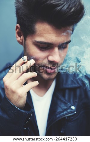 Close up portrait of charming fashionable man exhaling cigarette smoke while looking down, trendy attractive man blowing smoke out of his mouth standing on grey background, filtered image, blue flare