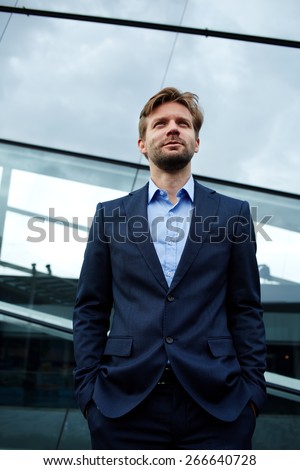 Low angle portrait of a handsome smiling businessman standing with his hands in his pockets in front of office building, carefree future vision concept