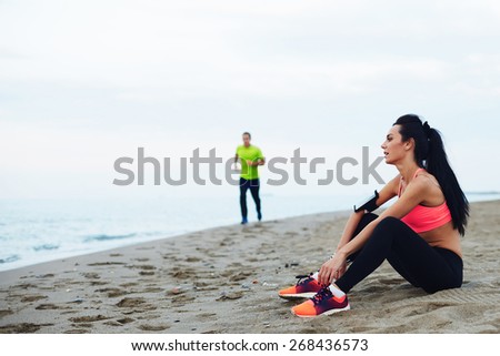 Athletic young woman in sportswear resting on the beach after exercise and looking at landscape, fitness man running along the beach looking at girl, sport people and active lifestyle