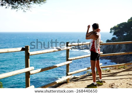 Side shot of handsome young runner stretching his arms before starting his run while standing on edge of a cliff with a wooden fence and enjoying ocean view from altitude
