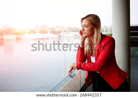 Portrait of pensive businesswoman looking out of an office balcony with beautiful seaport view on background, unhappy successful woman standing in exterior,blonde woman looking out of her window sadly