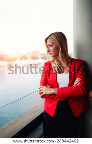Portrait of confident businesswoman looking out of an office balcony with beautiful seaport view on background, female executive with crossed arms thoughtfully looking away while having work break