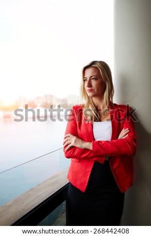 Portrait of confident businesswoman looking out of an office balcony with beautiful seaport view on background, female executive with crossed arms thoughtfully looking away while having work break
