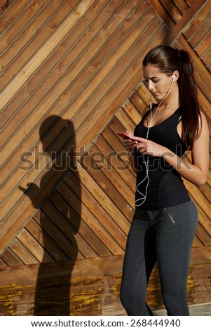 Attractive fit woman in sportswear listening to music with her headphones while training outdoors at beautiful sunny day, young female runner selecting music on cell phone while getting ready for run