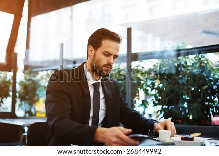 Portrait of businessman having breakfast sitting on beautiful terrace with plants, handsome well dressed man in suit using digital tablet for work sitting in cafe