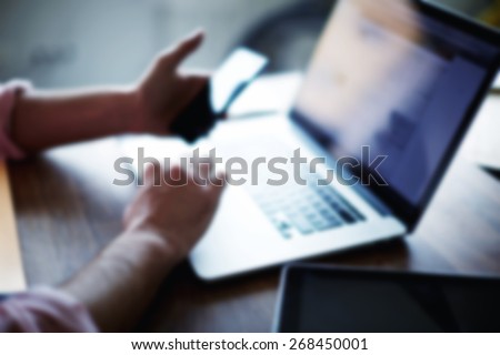 A cropped side shot of a young man working from home using smart phone and notebook computer, man\'s hands using smart phone in interior, man at his coworking place using technology blur effect