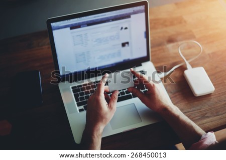 Rear view of male hands holding credit card using laptop at home with mobile phone and hard drive on the wooden table, data security, soft focus, flare sun light, cross process image