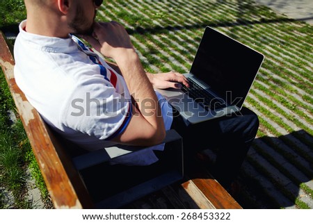 Modern young businessman freelancer working on his computer while sitting on city park bench, male student using notebook outdoors in urban setting while typing on keyboard, tourist working on laptop