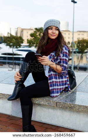 Gorgeous young woman using her smart phone while looking to the side outdoors, beautiful female student holding cellphone while sitting on the bench, hipster girl looking to someone in the park