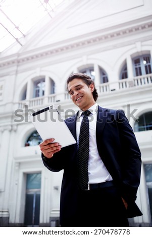 Man using digital tablet computer while standing in big light hall of railway station, smiling successful businessman on way to work reading news on his touch pad, happy executive receiving good news
