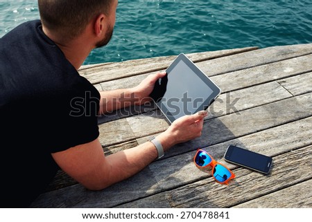 Young tourist man using digital tablet while lying on wooden jetty at marina port during his vacation holidays, man relaxing and enjoying outdoors while reading digital e-book on touch pad