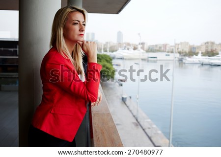 Portrait of pensive businesswoman looking out of an office balcony with beautiful seaport view on background, unhappy successful woman standing in exterior,blonde woman looking into the distance sadly