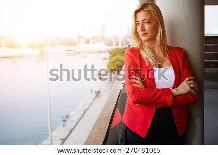 Portrait of confident businesswoman looking out of an office balcony with beautiful seaport view on background, female executive with crossed arms thoughtfully looking away, flare sunset light