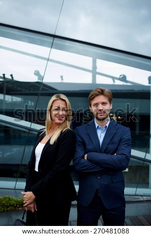 Two business colleagues standing against office building with brightly smile, experienced businessman standing with his female colleague smiling and looking to you, teamwork concept