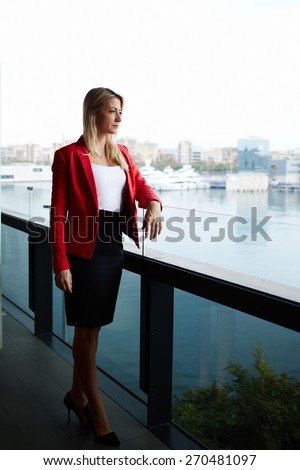 Elegant successful woman standing on the balcony of modern office building pensive looking away, rich well dressed woman standing in office exterior part with marina port view on background