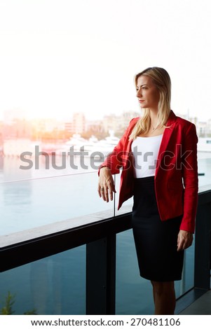 Elegant successful woman standing on the balcony of modern office building pensive looking away, rich well dressed woman standing in office exterior part with marina port view on background, flare sun