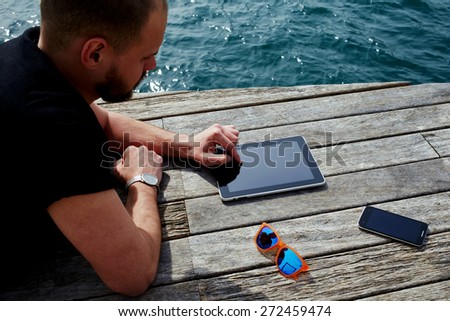 Young tourist man using digital tablet while lying on wooden jetty at marina port during his vacation holidays, man relaxing and enjoying outdoors while reading digital e-book on touch pad