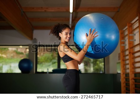 Young cheerful afro woman standing with fit ball in fitness center, attractive afro girl with curly hair smiling at gym
