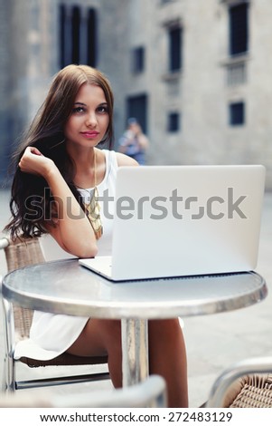 Pretty young woman in summer dress sitting at sidewalk cafe with open laptop computer, brunette hair female with open notebook next to her outside in coffee shop, tourist girl using her laptop in city