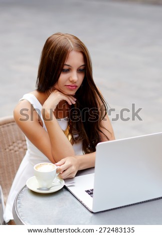 Beautiful young woman enjoying a cup of coffee with laptop open next to her, attractive female read digital book on computer while sitting outside in a street cafe, student girl use notebook outdoors