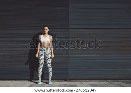 Young woman holding weights with hands by sides, sexy female look to you while taking break during her training outdoors against black copy space background for your text message, vintage pastel color
