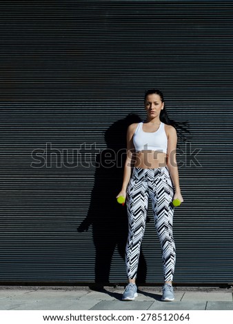 Sporty young woman holding weights with hands by sides against black wall background at sunset, sexy female look to you while working on her arms outdoors against copy space for your text message