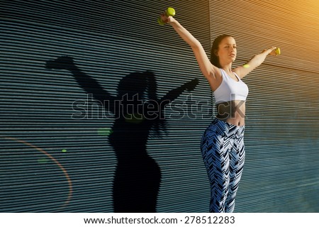 Athletic female with perfect figure getting her arms in great shape while lifting weights, attractive young woman using dumbbells to work out her arms while training outdoors on copy space background
